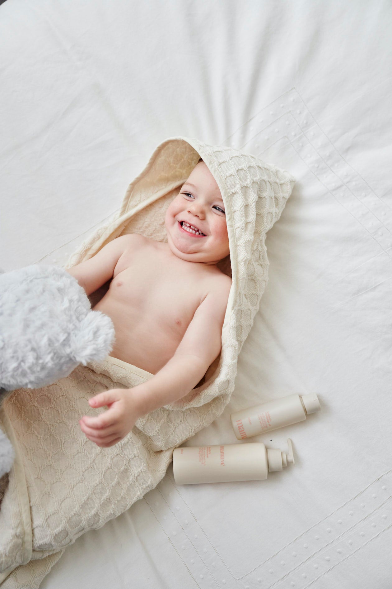 baby on bed with hooded towel on giggling with products beside and a blue teddy
